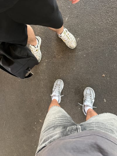 The image shows a top-down view of two people's legs and feet standing on a dark grey, textured surface, likely asphalt or a similar outdoor ground material, possibly in the city of Nîmes, France.

The person on the left is wearing black shorts, white socks, and light-colored athletic shoes with yellow accents and laces. A black bag is positioned next to their legs.

The person on the right is wearing grey denim shorts, white socks, and light grey athletic shoes with white laces.

The ground appears to be a common urban surface, consistent with outdoor walkways or public spaces in Nîmes. There are no other identifiable structures or landmarks visible in the frame. The lighting suggests it is daytime.