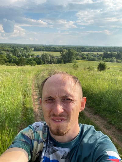 A selfie is taken by a man in his early 30s with light skin, receding hairline, and a short beard. He is wearing a camouflage t-shirt and standing on a dirt path in a grassy field. In the background, there are trees, houses, and a blue sky with clouds. The scene appears to be in the village of Nizhnii Bugrysh, Russia.