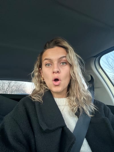A blonde woman with green eyes, wearing a black coat, seatbelt, and white sweater, looks surprised in a selfie. She's inside a car. The window shows a tree without leaves. She has on small earrings. The interior of the car includes a gray headliner and a light-colored panel above the window.