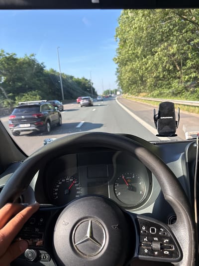 The image shows a first-person view from inside a vehicle driving on a highway. The steering wheel and dashboard are prominently visible in the foreground, with a hand resting on the steering wheel. A Grundig phone mount is attached to the dashboard. Various other cars are seen on the highway, suggesting traffic. Trees line the sides of the highway, indicating an outdoor, possibly rural or suburban, environment. The sky is clear and blue, suggesting daytime and fair weather. The speedometer indicates the vehicle is traveling at approximately 80 km/h. The license plate of a dark-colored SUV in front of the vehicle is visible. The location is the E19 highway in Mechelen, Belgium, as indicated by the context of the question.