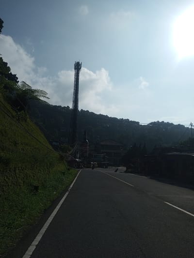 The image shows a road in what appears to be a rural or semi-rural area of Phouviang-Gnai, Laos. The road is asphalt and has white lane markings. A grassy slope rises on the left side of the road. Overhead power lines run along the slope. In the distance, there is a cell tower and a large bottle-shaped structure beside it. A few buildings are visible in the background, nestled amongst lush vegetation. There is at least one motorcycle on the road and a three-wheeled vehicle beside the bottle structure. The sky is light blue with some scattered clouds and the sun is bright.