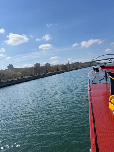 The image presents an exterior view from the starboard side of a vessel on the Danube–Black Sea Canal, near Medgidia, Romania. The foreground features the vessel's red, diamond-plate deck, which includes two black cylindrical mooring bitts, black lines or cables, and a partial yellow cylindrical object on the far right.
The canal's dark blue-green, rippled water occupies the mid-ground, extending towards both banks. On the left bank, a concrete embankment is visible, followed by a park-like area with numerous deciduous trees, mostly bare, indicative of early spring. Beyond the trees, several multi-story, light-colored residential buildings are positioned, signifying an urban environment.
In the distance, a prominent blue arch bridge spans the canal, connecting the distant banks. The terrain beyond the right bank and the bridge appears elevated and vegetated. The sky above is clear blue with scattered white cumulus clouds. No individuals are visible within the frame.