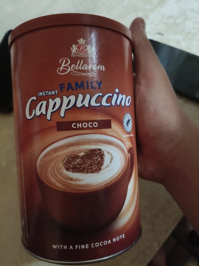 The image shows a hand holding a can of Bellarom Family Instant Cappuccino. The can is predominantly brown with white and blue lettering. The can displays the brand name "Bellarom" in a cursive font, "FAMILY" in blue block letters, and "INSTANT" above "Cappuccino" in large, stylized white letters. Below the cappuccino text, there is a smaller brown rectangular label with the word "CHOCO" in white letters. A circular emblem with a frog and the words "Rainforest Alliance" and "People & Nature" is also visible. At the bottom of the can, white text reads "WITH A FINE COCOA NOTE". The background is blurred and appears to be an indoor setting. There is no information in the image to indicate the location is Chornobaivka, Ukraine.