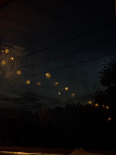 A dark, grainy image captures a nighttime scene, possibly from a window. In the foreground, a blurred wooden surface runs horizontally across the bottom. The background depicts a deep blue-black sky with several horizontal lines that could be power lines. Scattered across the sky are blurry, glowing yellow orbs, likely reflections or lights. Towards the middle of the frame, a faint crescent moon is visible. Silhouetted against the sky is a dark line of trees and foliage. To the right, the dark silhouette of a tree with visible branches stands out. The overall impression is one of a dimly lit, possibly rural or suburban, outdoor environment at night. There is no specific indication of the city of Jāņupe, Latvia.