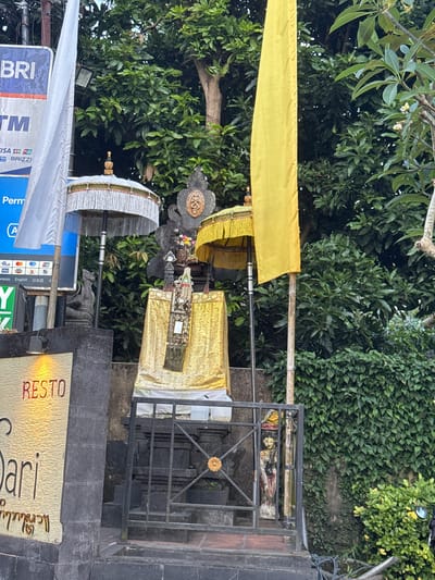 The scene is a street in Mas, Indonesia, featuring a religious shrine or offering place. The shrine is adorned with a yellow cloth and an umbrella. In the background is a wall or gate made of black metal and stone, with what appears to be a framed image behind the bars. Nearby, there are signs and advertisements, one displaying ATM information and another advertising a restaurant labeled "Resto" in red. Lush green trees and shrubs form the backdrop, suggesting a tropical climate. A large BRI sign is visible.