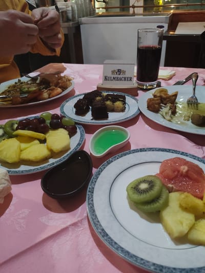 The scene is a table laden with food, likely in a restaurant or dining hall in Bayreuth, Germany, as indicated by the "Kulmbacher" drink container visible near a glass filled with a dark liquid (possibly red wine). The table is covered with a pink tablecloth. Various plates are scattered across the table: one holds fried food, another displays slices of grapefruit, kiwi, and pineapple. A third plate contains brownie-like pastries, and a fourth showcases more fruit like grapes, pineapple, and kiwi. Small containers of sauces or jellies are also present. A person, partially visible on the left side of the image, is interacting with something out of frame, possibly preparing to eat.