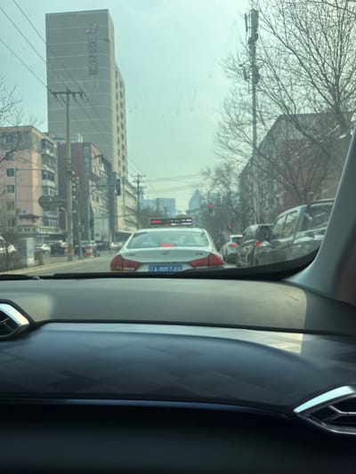 The image presents a daytime street scene viewed from the interior of a vehicle, looking forward through the windshield. The foreground shows the dark dashboard of the observer's car, which features a subtle geometric pattern and a chrome-accented air vent on the right side.

Immediately ahead, a white sedan with a Chinese license plate displaying "A MM74R" is stopped in traffic, its red brake lights illuminated. A red LED display unit is visible on the roof of this sedan. Further down the multi-lane road, additional vehicles are present, including a dark SUV on the right, all appearing stopped or moving slowly. Distant red traffic lights are visible.

On the left side of the road, a prominent, light-colored, multi-story building features vertical Chinese characters "锦江之星" (Jinjiang Inn) on its facade. Next to this building are several smaller, older structures, likely residential or commercial. Multiple power poles with overhead electrical wires line the street. On the right side of the road, bare-branched trees stand, and a tall cellular tower equipped with antennas is visible. The sky is a light, hazy grey.

The scene is located in Huanggu District, Shenyang, China, identified by the Jinjiang Inn signage and the "A" prefix on the license plate, which denotes Shenyang within Liaoning Province.