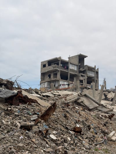 A destroyed building stands amidst rubble under a cloudy sky. The multi-story structure is heavily damaged, revealing exposed rooms. Debris consisting of concrete, metal, and various materials piles up around the building's base, reaching a significant height. The scene conveys devastation and destruction.