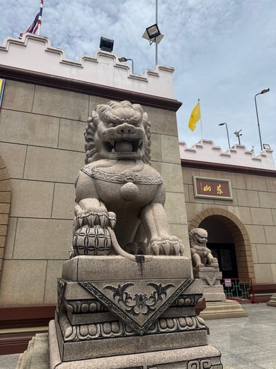 In a medium shot, two stone lion statues stand guard in front of a building with a crenellated pink parapet. The foreground statue, a large, intricately carved lion with a fierce expression, is positioned on a stone pedestal adorned with black floral and geometric patterns. The lion is seated, with its front paws resting on a decorative spherical object. To the right and further back, a smaller, similar lion statue sits on a matching pedestal.
The building behind the statues has a light beige, stone-block facade. Above the arched entrance, a rectangular sign with gold lettering on a blue background is mounted. A yellow flag flies from a pole on the roofline. The sky is overcast with scattered clouds. The overall scene appears to be an outdoor location in the city of มาบฟักทอง, Thailand, likely a temple or cultural site given the architectural style and statues.