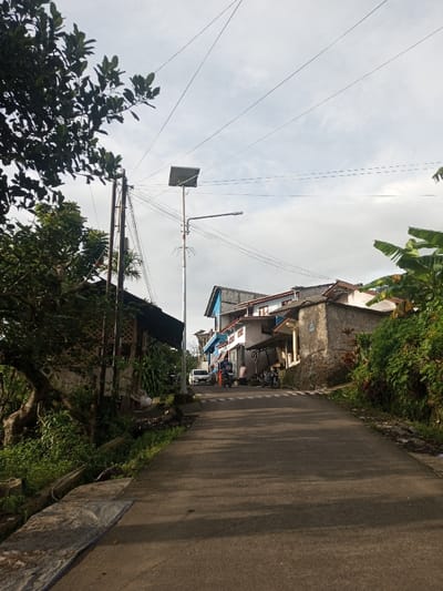 The image displays a residential street or pathway sloping upwards in what appears to be a semi-urban or rural setting, identified as Yusipang, Bhutan. The paved road dominates the foreground and midground, with both sides flanked by houses and lush vegetation.
On the left side, dense green trees and foliage partially obscure a building. A tall utility pole stands prominently, adorned with multiple overhead wires, a solar panel at its apex, and a street light. Another utility pole is visible behind it, also supporting numerous electrical wires that crisscross the sky.
Further up the road, several multi-story houses are visible, constructed with concrete or stucco and featuring various roof styles, some with blue-painted elements. In the distance, a few individuals are discernible near parked motorcycles and a white car, indicating some human activity. The sky above is partly cloudy, and the lighting suggests it is late morning or early afternoon, casting shadows across parts of the road. The environment is rich with greenery, including banana leaves visible on the right. No discernible text is visible.