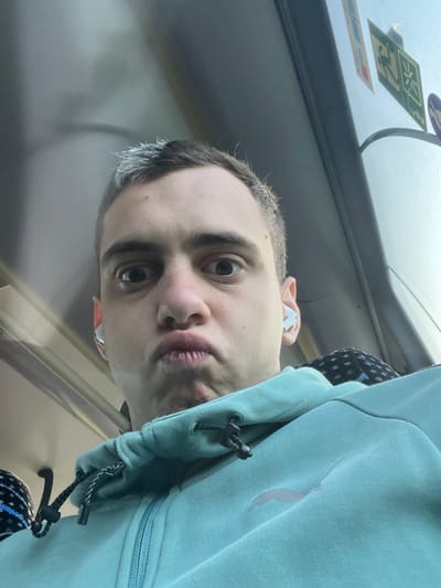 The image shows a close-up, selfie-style shot of a young Caucasian male from the chest up. He is wearing a teal hooded jacket with a zipper and drawstrings, and has white earbuds in his ears. His hair is dark with some grey streaks at the front. He is making a "fish face" expression, with his lips pursed. He has wide eyes and a slight grimace. The background appears to be the interior of a vehicle, possibly a bus or train, with windows and some stickers visible. The lighting suggests it is daytime. The image does not contain any information that could be used to determine the specific location within the city of Maardu, Estonia.
