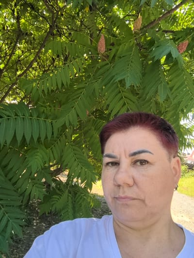 A middle-aged woman with short, dark red hair and light-colored eyebrows poses for a selfie outdoors. She is wearing a white t-shirt and has a piercing above her left eyebrow and a hoop earring in her right ear. She is positioned in front of a large tree with abundant green leaves and some cone-shaped seed clusters. The background suggests a rural or suburban setting with a dirt path, grassy areas, and a glimpse of houses in the distance. The lighting indicates it is daytime. The overall scene is relaxed and informal.