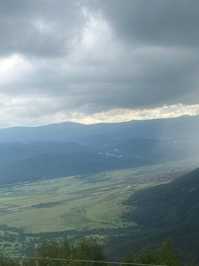 The image displays a sprawling natural landscape captured from an elevated perspective, oriented with the horizon running vertically down the middle. When viewed horizontally (rotated 90 degrees clockwise), the scene reveals a vast sky dominated by a mix of grey and bright white clouds, suggesting a partly cloudy or overcast day with intermittent sunlight breaking through.
Below the sky, a long, dark, and rugged mountain range extends across the mid-ground, its slopes densely covered in forests. The light highlights certain contours of the mountains, indicating their scale and texture.
Further down, in the foreground (when rotated), lies a wide valley or plain. This area is characterized by a patchwork of lush green agricultural fields, interspersed with darker patches of trees and what appear to be small clusters of buildings, likely forming a village or small settlement in the distance along the base of the mountains. Faint, lighter lines within the valley could indicate the presence of rivers or streams.
On the far right edge of the image (when rotated), a thin, white vertical line is visible, which appears to be a power line or cable, indicating the presence of infrastructure in the remote landscape.
The time of day appears to be daytime, with diffused light illuminating the scene, creating a serene and expansive atmosphere. The landscape is typical of a mountainous region with agricultural valleys, consistent with the provided location context of Karnare, Bulgaria. No specific human activity or visible text is present.
