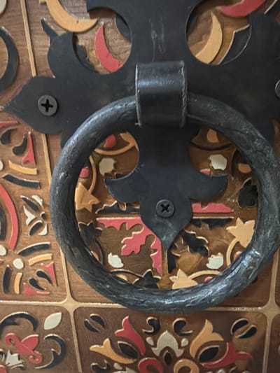 The image shows a close-up of a decorative wooden surface with a metal ring handle. The wood has an inlaid design of various shapes and colors, including red, yellow, white, and black. The metal handle is dark and appears to be wrought iron, attached to the wooden surface by black metal embellishments. There are visible screws holding the metal embellishments in place. The overall impression is of ornate, possibly antique, craftsmanship. No people, actions, or interactions are visible. The location is not discernible from the image.