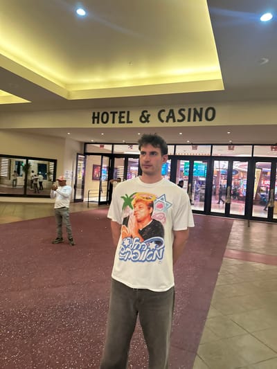 The image depicts a man standing in what appears to be the entrance of a hotel and casino. The words "HOTEL & CASINO" are prominently displayed on a column to his left. The interior of the casino is visible through glass doors behind him, showcasing various gaming machines and people. The man is wearing a white T-shirt with a graphic design and grey pants. Another person is seen walking behind the man. The scene is located in Hollywood Seminole Reservation, United States of America.