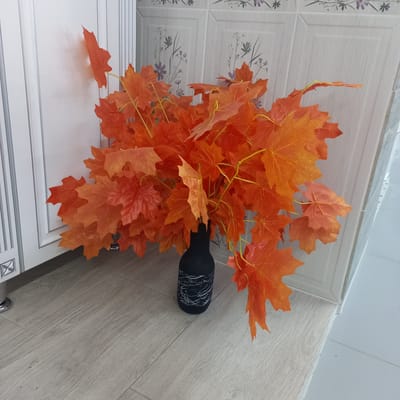 A high-angle, close-up shot captures a bouquet of vibrant orange artificial maple leaves arranged in a black vase with white swirling patterns. The vase sits on a light-colored, wood-effect floor, with the leaves spilling out and partially resting on the edge of a white cabinet. The background features a wall with light-colored, textured tiles adorned with a subtle floral pattern of grey stems and small purple flowers. The lighting appears to be from an overhead source, casting soft shadows. The overall scene suggests an indoor setting, possibly a home or a decorative display, with an autumnal theme. There is no visible text in the image.