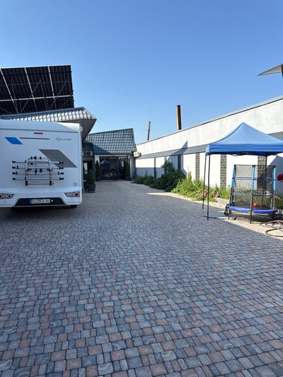 In Parakar, Armenia, a white RV is parked on a paved driveway. To the left, solar panels are installed on a roof. In the background, a building with a dark-tiled roof and a large glass entrance is visible. Adjacent to the building, a blue canopy shelters a trampoline with a small orange ball on it. The scene is outdoors on a clear, sunny day, with lush greenery and flowering plants lining the driveway.