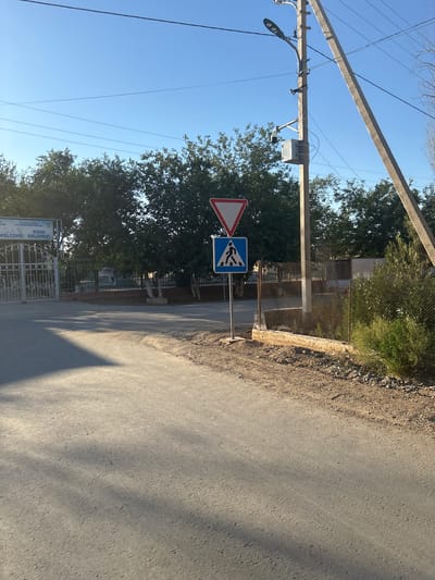 The image depicts a street scene in Nukus, Uzbekistan, likely on a sunny day given the clear blue sky and strong shadows. In the foreground, a paved road curves to the right, with a gravel and grassy shoulder to the right. Tall trees with dense green foliage line the background. On the left side of the road, a white and silver gate with intricate metalwork is visible, with a sign above it that reads "WELCOME! KELDINIZ!". This suggests the entrance to a facility, possibly an educational institution or a public building.

To the right of the gate, a prominent utility pole stands, equipped with a streetlight and a security camera. Further down the road, a pedestrian crossing sign is displayed on a pole, with a white triangle with a red border above a blue square containing a white pedestrian icon. This indicates a designated crossing area. A chain-link fence runs along the right side of the road, bordering an area with more vegetation. The overall environment suggests a relatively quiet street, possibly on the outskirts of the city or in a residential area.