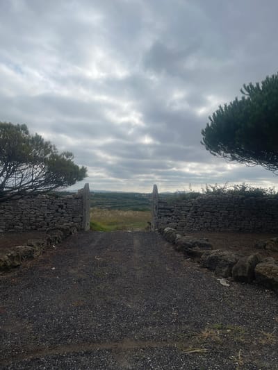 This is a photograph of a stone gateway leading into a landscape under a cloudy sky. The gateway is flanked by two low, rustic stone walls, appearing to be made of irregularly shaped rocks. The ground in front of the gateway is covered in dark gravel. On the left side of the gateway, a tree with sparse foliage extends over the stone wall. On the right side, the branches of a conifer tree are visible. Beyond the gateway, the landscape opens up to rolling hills covered in vegetation, with distant structures visible on the horizon. The overall impression is one of a rural or natural setting, possibly a private property or an entrance to a park or natural reserve. The location is specified as São João das Lampas, Portugal.