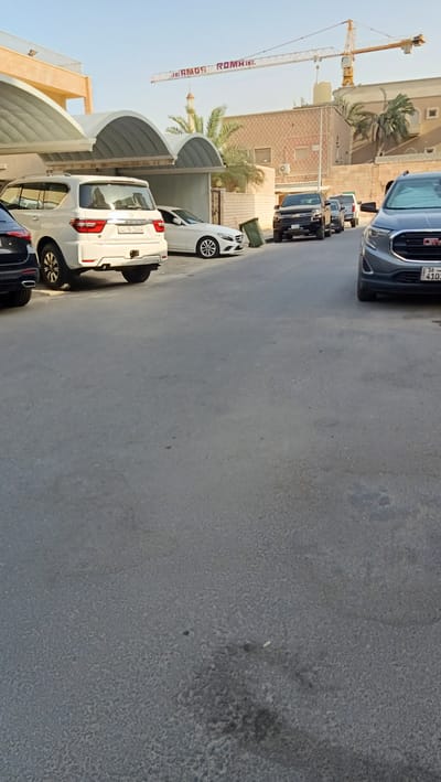The image shows a street scene in Rumaithiya, Kuwait. Several cars are parked on the side of the road. In the background, there is a building with a crane visible above it. The sky is clear and the sun is shining. The overall scene appears to be a typical day in the city.