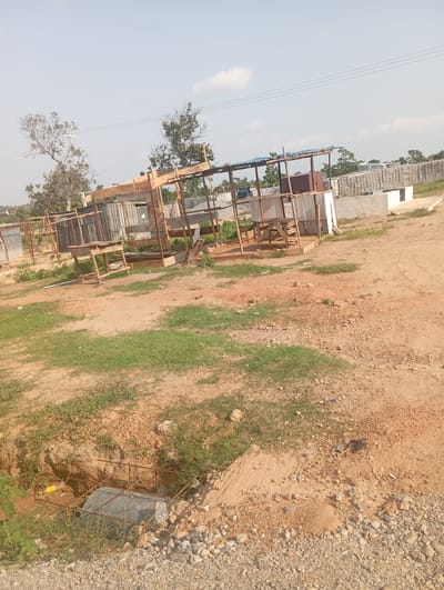 The image depicts an outdoor scene in Tasha, Nigeria, characterized by a dirt road and a construction site. In the foreground, a partially dug trench with rebar reinforcement and a cylindrical object covered in plastic is visible. The surrounding area is covered with sparse vegetation and loose gravel.
Further back, a rudimentary open-air structure is under construction, featuring a wooden frame and corrugated metal roofing. Several wooden tables and benches are present within and around this structure. Adjacent to it, stacks of building blocks are piled high, indicating ongoing construction or material storage. Other simple structures, possibly temporary shelters or market stalls, with metal sheeting are also scattered throughout the area.
In the distance, trees with bare branches suggest a dry season. Power lines are visible stretching across the sky, supported by utility poles. The overall environment appears to be an undeveloped or semi-developed area, likely a construction zone or an informal marketplace on the outskirts of Tasha. There are no visible people in the image.