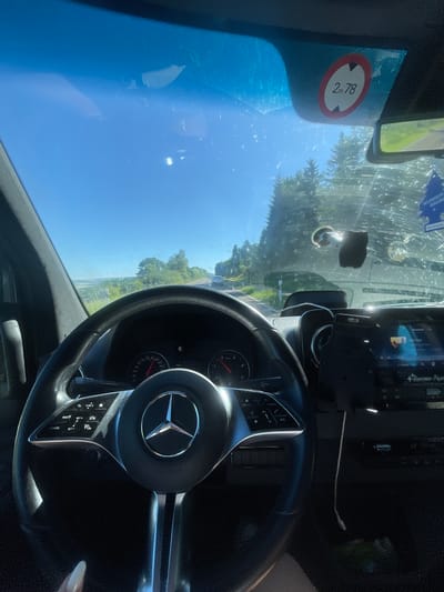 The image depicts the interior of a Mercedes-Benz car, specifically the steering wheel and dashboard, with a view through the windshield. The sky is blue, and trees and vegetation are visible outside, suggesting a rural or suburban setting. A sticker on the windshield reads "2m78", possibly indicating a height restriction or toll information. The location is identified as Blankenheim, Germany, based on the sticker's format common in European road signage. The car appears to be stationary, as there is no visible motion blur. The lighting is bright and sunny, casting shadows within the vehicle. A phone mount and a small air freshener are visible attached to the windshield. A USB cable leads to the device from a spot in the central console.