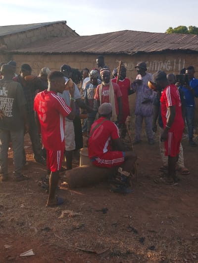 In a rustic setting, a group of people gathers outside a weathered building. A man in a red and white soccer uniform drinks from a gourd-like cup, while another, similarly dressed, sits on a log next to a metal container. Other individuals stand around them, some wearing traditional headwear and clothing. The ground is dirt, and the atmosphere seems to be one of community and tradition.