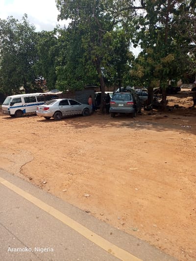 The image shows a dirt parking lot with three cars and several people in the town of Okinni, Nigeria, at coordinates latitude: 7.832, longitude: 4.51. The parking lot is bordered by a paved road with a yellow dividing line on the right side of the image. On the left side of the parking lot, there is a white van. In the center of the image, there are two cars, a silver sedan and a grey SUV, which appear to be parked. There are five individuals in the parking lot, all of whom are standing or walking. They are all facing away from the camera. The lot is mostly dirt, with a few scattered leaves and pieces of trash. There are trees on the left, right, and in the background. The sky is overcast.