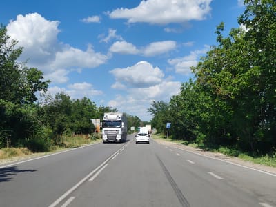 The image depicts a daytime scene on a two-lane asphalt road, likely in or near the city of Yasen, Bulgaria, based on the provided information. The sky is blue with scattered white cumulus clouds. Dense green trees line both sides of the road, with some patches of dry grass and vegetation on the shoulders.
In the foreground, the road is marked with double white lines. A white passenger car is traveling in the right lane, with its rear visible. Ahead of the car, in the left lane, is a large white semi-trailer truck. The truck has a white cab with the DAF logo and "SUPER S..." visible on the front. The trailer appears to be a box trailer. Further ahead, more vehicles, including another white truck and possibly a van, are visible in the distance.
On the right side of the road, a blue and white traffic sign is present. The overall impression is a highway or a main road with moderate traffic on a clear day. No people are explicitly visible.