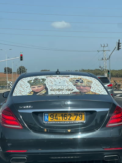 The image displays the rear of a dark grey Mercedes-Benz S 500e sedan, stationary at a traffic light in Kfar Yona, Israel. The vehicle features an Israeli license plate, number 94-162-79, with "ת.א.מ. מרכז שירות לרכב בע"מ 03-6134134" (T.A.M. Car Service Center Ltd.) printed below the numbers. The entire rear window is covered by a patterned decal depicting two stylized portraits: a woman wearing a multi-jeweled crown, identified as "QUEEN TAMAR," on the left, and a man wearing a golden crown with a blue Star of David on the right. The decal includes the English text "THE ONE," the website "www.SHARSHAR.CO.IL," a QR code, and the Hebrew phrase "אין כמו בבית" (There's no place like home).
In the background, a traffic light on the left is illuminated red, while another further to the right appears green. Streetlights, utility poles with power lines, and distant vegetation are visible. The sky is clear blue with one small white cloud. A light-colored SUV is partially visible behind and to the right of the Mercedes, with an individual on a small motorized scooter or bicycle in the immediate vicinity. The perspective is from a vehicle directly behind the Mercedes-Benz.