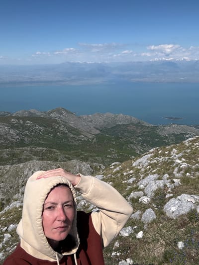 A person wearing a two-toned hoodie is lying on a rocky, grassy surface. The person's head is encased in the hoodie. In the background, there is a mountainous landscape extending to a body of water and the blue sky. The photo appears to be taken from a high altitude, possibly in or near Mali Mikulići, Montenegro.