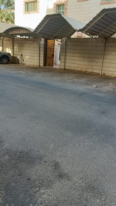 The image shows a street view in Rumaithiya, Kuwait. In the background, there is a building with beige and white brickwork, featuring light-colored windows with visible frames. In front of the building, a wall constructed from large, light-colored concrete blocks runs horizontally across the frame. Above this wall, a series of metal awnings with corrugated metal roofs are installed, providing shade over a walkway or parking area. An ornate golden gate with intricate scrollwork is visible in the center, slightly ajar, revealing a glimpse of what appears to be an interior. To the left of the gate, a portion of a dark-colored vehicle is parked. The foreground is dominated by an asphalt road, showing a textured surface with some wear and tear. Scattered on the road and sidewalk area are a few small pieces of debris, including what appears to be a crumpled piece of paper. The lighting suggests it is daytime.