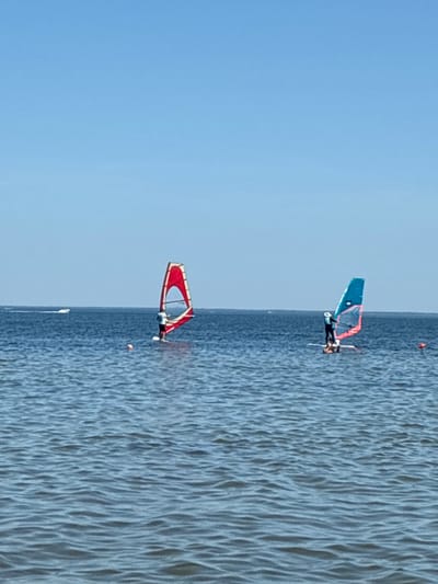 Two individuals are windsurfing on a calm, blue body of water under a clear sky. The individual on the left is on a board with a red and yellow sail, while the individual on the right is on a board with a blue and pink sail. In the background, a small white boat is visible. The image does not provide any information about the city of Rewa, Poland.