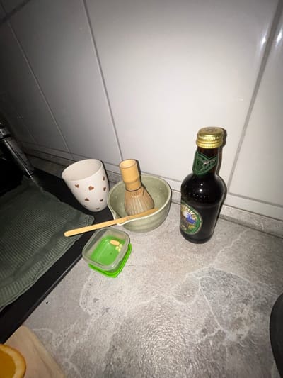 The scene depicts a countertop arrangement in an indoor setting, likely a kitchen, located in Hagen, Germany. The countertop surface is grey with a mottled or textured pattern, backed by a white tiled wall with visible grout lines.

From left to right:
On the far left, a portion of a chrome-finished faucet is visible. Adjacent to it, on a dark, flat surface, a dark green, ribbed textile item, possibly a towel, is neatly folded. Partially visible in the bottom-left corner is a slice of an orange resting on a light-colored wooden cutting board.

Centrally positioned on the countertop is a white ceramic mug adorned with multiple small, reddish-brown heart patterns. To its right, a greenish-grey ceramic bowl contains a light brown bamboo matcha whisk and a light brown bamboo matcha scoop. In front of this bowl, slightly to the left, rests a small, rectangular, clear plastic container with a bright green lid. Inside this container, approximately four pills, yellow and white in color, are visible.

To the right of the bowl, a dark brown glass bottle stands upright. It features a gold-colored twist-off cap and a green label, which includes an unidentifiable graphic (appearing as a landscape) and unreadable text. The scene is illuminated by an overhead light source, casting soft shadows. No people are present, and no actions are in progress.
