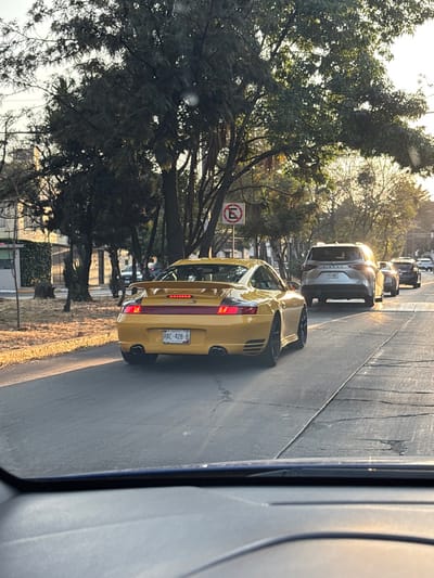 A gold car, possibly a Porsche, is seen from behind while driving on a road with other vehicles. A "No Parking" sign is visible on the left amidst trees. The photo is taken from inside a vehicle, showing part of the interior. The scene is bathed in sunlight, suggesting it's daytime. The car has a license plate that reads "RAC-428-B".