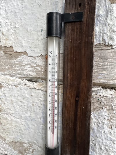 The image shows a thermometer attached to a weathered wooden post. The thermometer displays a temperature of approximately 5.7 degrees Celsius. The background consists of a textured, peeling white wall and a rustic wooden plank. The scene appears to be outdoors, possibly in a rural setting or an older part of a town. No people or specific landmarks are visible. The thermometer is the primary subject, indicating a cool ambient temperature.