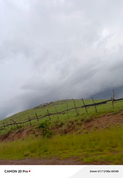 The image depicts a landscape scene, likely a hillside, under an overcast sky. A wooden fence runs diagonally across the grassy slope. In the foreground, there is an area of brown earth and vegetation. The bottom of the image displays text identifying the device as "CAMON 20 Pro" and camera settings: "24mm f/1.7 1/150s ISO50". While the text does not explicitly mention Nguroje, Nigeria, the overall environment suggests a rural or natural setting that could be found in such a location. There are no people or specific actions visible in the scene.