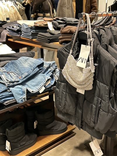 The image provides a close-up view of a clothing display within a Zara retail store in Chalkida, Greece. The scene is well-stocked with various garments and accessories, suggesting a shopping environment.
In the foreground and center, a shimmering silver, rhinestone-embellished top or small bag hangs prominently from a rack, with a white price tag clearly displaying the "ZARA" logo. Immediately behind this sparkling item and to its right, several black puffer vests or jackets are neatly arranged on hangers. Another "ZARA" tag is visible on one of these black garments towards the upper right side of the frame.
Below the hanging black puffer items, a shelf holds folded blue denim jeans, indicating a section for casual wear. To the bottom right, a pair of black puffer-style boots or slippers is displayed on a wooden shelf, with a "ZARA" tag attached to one of the shoes. Further to the left and in the background, more clothing items, including brown jackets and dark pants, are visible on racks and shelves, contributing to the typical cluttered yet organized appearance of a busy clothing store.
The setting is indoors with artificial lighting, and there are no people visible. The focus is entirely on the merchandise. The visible text "ZARA" appears multiple times on price tags, confirming the brand of the items.
