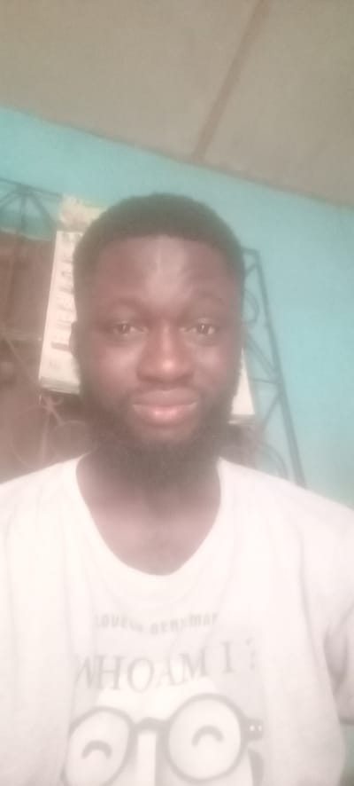 A medium-shot photograph depicts a dark-skinned male with short, dark hair and a full beard, facing the camera with a slight smile. He is wearing a light-colored t-shirt with visible text and a graphic near the bottom. The background consists of a light blue wall, a portion of a calendar or document hanging on the wall, and the corner of a metal frame. It is not possible to verify the location of the image within the city of Bende, Nigeria from the provided image.