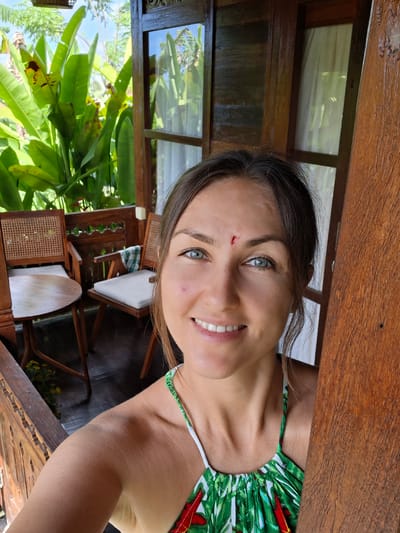 A woman is posing for a selfie, taken in Lodtunduh, Indonesia. She has brown hair and blue eyes and is wearing a green and white tropical-print top. There is a small cut on her forehead. The background includes a wooden structure with a window and lush green vegetation. Additionally, there are wooden chairs with white cushions visible. The perspective is slightly tilted.