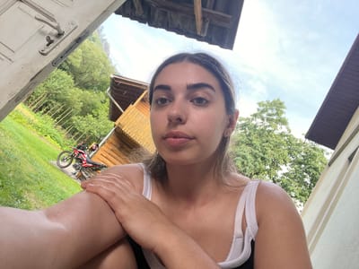 The media file is an upside-down, close-up selfie of a young woman. She is wearing a white tank top and her arms are crossed in front of her chest. Her face is the central focus, with her eyes looking directly at the camera. Her lips are slightly parted.
The background suggests an outdoor setting, possibly a rural or semi-rural area. To the right of the woman, a wooden structure, possibly a shed or small building, is visible. A red and white dirt bike is parked near this structure. Further in the background, lush green trees and grass indicate it is likely daytime and the weather is fair. The sky is visible above the trees, appearing bright and possibly sunny.
To the left of the woman, a section of a cream-colored wall or structure is present, with horizontal slats. At the bottom of the frame, a portion of a weathered, pale-colored door is visible, including a door handle and lock mechanism.
The overall impression is a casual, candid moment captured outdoors, perhaps in a backyard or near a dwelling. The upside-down orientation of the image adds an unusual perspective. There is no visible text in the media file.