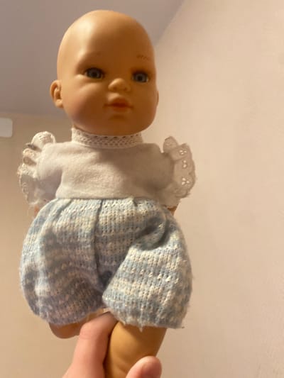 A bald baby doll is held by a hand, with the doll wearing a white top and blue knit shorts. The background is a plain, light-colored wall. This image was captured indoors in the city of Afanasyevo, Russia.