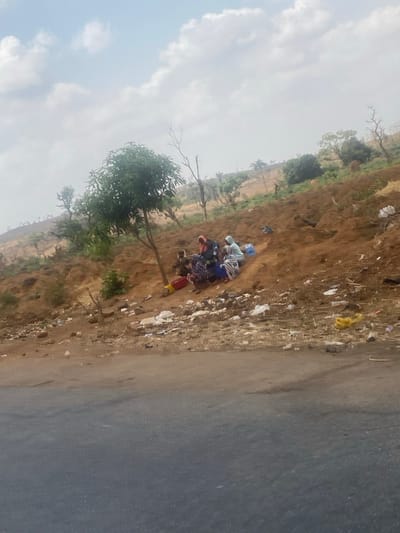 The image depicts an outdoor scene in Nandu, Nigeria, featuring approximately seven individuals, primarily women, seated on a reddish-brown dirt embankment adjacent to a paved road. The group is clustered near the base of a small, green-leafed tree. Individuals wear various garments, including head coverings, with visible colors such as light blue, red, and dark patterns. A red bucket or bowl and a blue container are present near the seated figures. The immediate area of the embankment is littered with white plastic bags, yellow plastic debris, and other assorted waste. Beyond the embankment, the landscape consists of sloping hills with sparse vegetation, including several bare or dry trees, extending towards a hazy background under an overcast sky. The dark gray paved road occupies the lower portion of the frame.