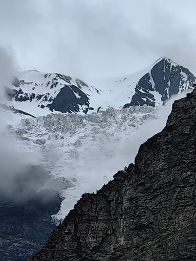 A dramatic, low-angle, vertical shot captures a majestic, snow-capped mountain range under a cloudy sky. The foreground is dominated by a steep, dark, rocky cliff face on the right, textured with intricate patterns of erosion and shadow. Beyond the cliff, a vast, jagged glacier spills down from the mountain peaks, appearing as a turbulent river of ice. The glacier is heavily crevassed, with a rough, uneven surface.
Behind the glacier, the imposing mountains rise, their upper reaches covered in a thick blanket of snow. Dark, rocky outcrops punctuate the white slopes, creating sharp contrasts. The sky is overcast with heavy, grey clouds, obscuring any direct sunlight and contributing to a cool, somber atmosphere. The overall scene conveys a sense of raw, untamed natural power and the formidable scale of the mountain environment. Two tiny figures are visible on the dark rock in the far right of the image, providing a sense of scale against the colossal landscape.
The image appears to have been taken during daylight hours, given the visibility, but the heavy cloud cover suggests overcast weather, possibly cold and damp. No text is visible in the image. The setting is clearly a mountainous region with a significant glacial presence, consistent with a location like Gemur, India, known for its high-altitude terrain and glaciers.