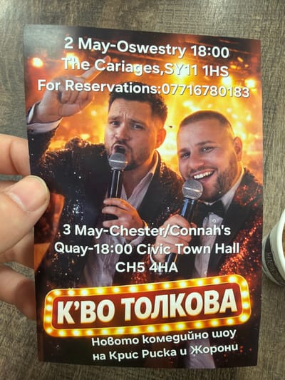 A glossy paper flyer is held in the upper left quadrant by a human hand, primarily the thumb, against a textured brown surface. The flyer advertises a comedy show, featuring two adult males depicted from the mid-chest up, both wearing dark, sequined jackets and holding silver microphones against an orange and yellow bokeh-lit background. The man on the left, with short dark hair and a beard, wears a white shirt beneath his jacket, holds a microphone near his mouth, and raises his right arm with his index finger pointing upwards. The man on the right, also with short dark hair and a beard, wears a white shirt and black bow tie under his jacket, holds a microphone, and displays a broad, open-mouthed smile.
White text overlays the image, detailing two event dates: "2 May-Oswestry 18:00 The Cariages,SY11 1HS" with "For Reservations:07716780183" directly below it. Further down, "3 May-Chester/Connah's Quay-18:00 Civic Town Hall CH5 4HA" specifies the second event, identifying "Civic Town Hall, CH5 4HA" as the location in Connah's Quay, United Kingdom. At the bottom of the flyer, a prominent red and yellow illuminated sign-like graphic displays the show title in Cyrillic: "К'ВО ТОЛКОВА" (K'VO TOLKOVA), with smaller Cyrillic text below it reading "Новото комедийно шоу на Крис Руска и Жорони" (The new comedy show of Chris Ruska and Zhoroni). A white and black patterned cup is partially visible on the right edge of the frame, resting on the same brown surface as the flyer.