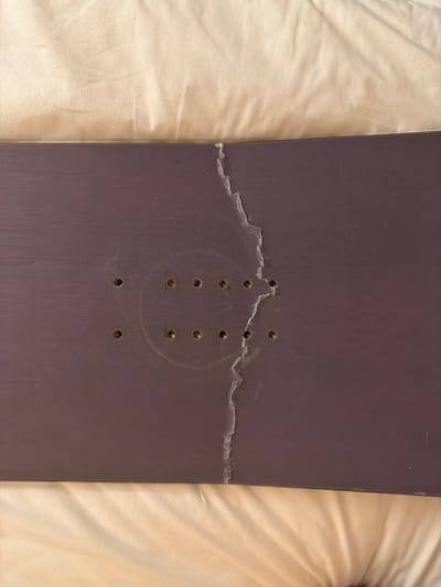 A damaged snowboard base, dark purplish-brown in color, is depicted in a top-down, close-up view, resting on a light-beige, wrinkled fabric surface. A prominent, irregular, jagged vertical crack with exposed white material along its edges extends through the center-right section of the snowboard. To the left of this crack, a pattern of twelve small, circular, metallic binding insert holes is arranged in three horizontal rows of four, centered within a faint circular impression on the board's surface. No individuals, specific actions, or environmental elements are visible to confirm the scene's location within the city of Kutchan, Japan.