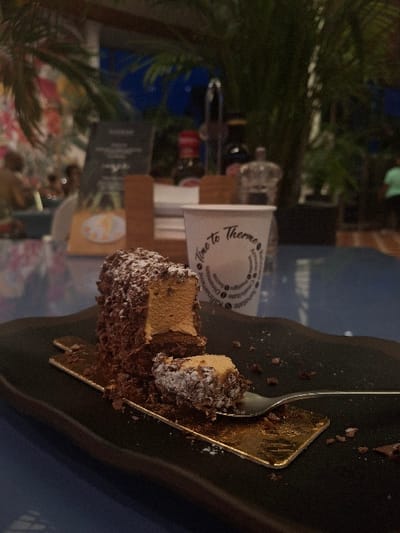 The scene depicts a partially consumed chocolate dessert and a beverage cup on a table within an indoor dining establishment in Săftica, Romania, specifically implied to be near Therme București due to cup branding. In the foreground, a dark, oblong plate holds a rectangular chocolate bar dessert, heavily coated in chocolate shavings and lightly dusted with powdered sugar. A segment of the dessert has been cut, revealing a light brown, creamy interior. A small, sugar-dusted piece of the dessert rests on a silver-colored fork, which is positioned on a small, gold-colored foil wrapper beneath the main dessert. Chocolate crumbs are scattered across the plate and foil. To the right, slightly behind the dessert, is a white disposable cup with the text "Time to Therme" prominently displayed, along with other circular text. The table surface is blue. In the blurred background, lush green foliage is visible. On the table, a wooden caddy holds small condiment bottles, including one with a red cap, and a clear shaker. A menu board is also present. Distant, blurred figures of people and colorful wall decor are faintly discernible, indicating a public setting. The overall lighting is dim.