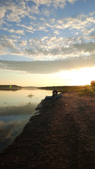 The image depicts a riverside scene at golden hour in Шолья, Russia, characterized by low-angle sunlight from the right. The upper sky displays a clear blue field populated by numerous scattered altocumulus clouds, with a significant sun glare emanating from the upper right quadrant.
A wide, calm body of water, likely a river, spans the mid-ground, reflecting the sky and clouds. The immediate foreground features a dark, sandy and pebbly shoreline, exhibiting subtle ripples and tire tracks across its surface.
Two individuals are present on the shore. On the left, closer to the viewer, a person dressed in camouflage-patterned clothing stands facing the water, holding a thin stick. Further to the right, another person is depicted in silhouette, bending over near a grey inflatable boat that has been pulled onto the sand.
The distant horizon beyond the water shows a low-lying landmass with a dark silhouette of vegetation, against which a faint structure, possibly a power line tower, is visible on the left. A distinct green lens flare is present on the left side of the image, superimposed over the distant land.