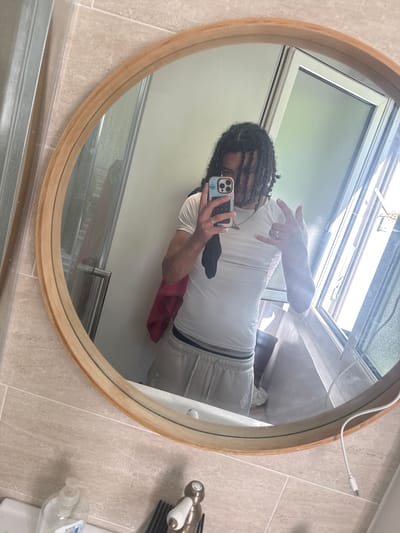 This is a first-person, mirrored selfie taken in a bathroom. The subject, a person with dreadlocks, is wearing a white t-shirt and light-colored athletic shorts with a dark stripe and branding on the waistband. They are holding a smartphone in their left hand, which is pointed towards the mirror, capturing the reflection. Their right hand is raised and making a hand gesture. A silver ring is visible on their right hand. The background shows a tiled wall and a round wooden-framed mirror. A window is visible in the reflection, suggesting natural light. A white charging cable is also visible extending from the top right of the mirror frame. To the right of the mirror, on the counter, a faucet and what appears to be a comb are visible, along with a bottle of hand sanitizer. The overall setting appears to be a residential bathroom. The image does not provide explicit cues for time of day or weather, but the presence of natural light from the window suggests it is daytime. There is no visible text, other than potential branding on the shorts, which is not clearly legible. The emotions conveyed are neutral, typical of a casual selfie.