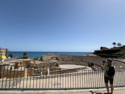 The image depicts the Roman Amphitheatre of Tarragona, a historical site located in Tarragona, Spain. The amphitheatre, constructed from stone, is partially ruined, revealing its tiered seating and central arena. In the foreground, a person wearing a green t-shirt, shorts, a hat, and a backpack is standing behind a metal fence, holding up a phone to take a picture of the amphitheatre. The person is facing away from the camera, looking towards the historical structure. Behind the amphitheatre, the Mediterranean Sea stretches out under a clear blue sky, with several cargo ships visible on the horizon. To the left of the amphitheatre, there is a modern building and a yellow container. The overall scene suggests a bright, sunny day with a tourist exploring the ancient ruins.